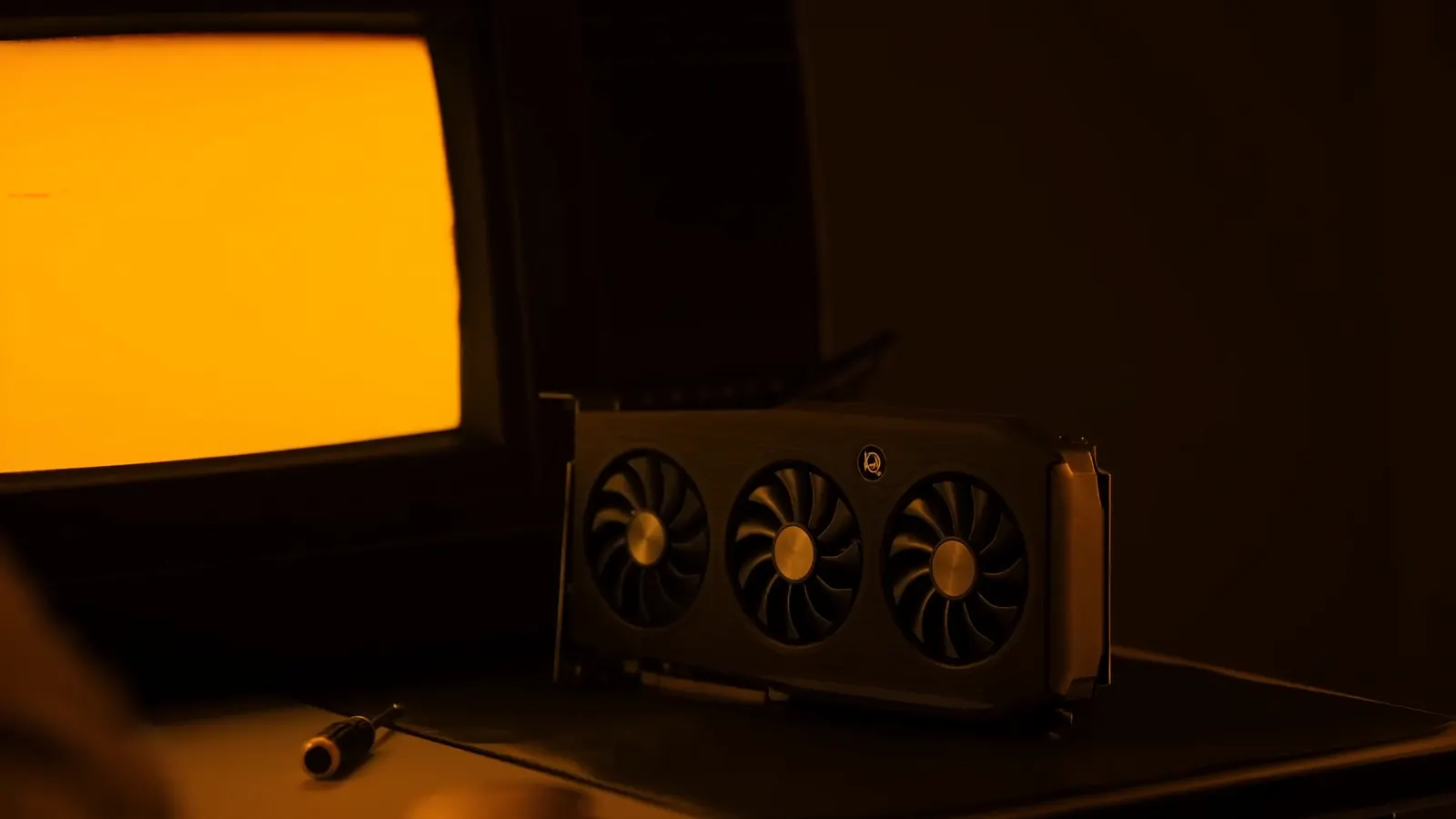 A triple-fan graphics card on a workbench, lit by the amber glow of a CRT monitor